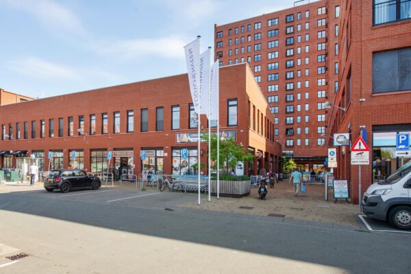 Moderne rode bakstenen gebouwen aan het Harriet Freezerhof met winkels, vlaggenmasten en geparkeerde auto's op de voorgrond.
